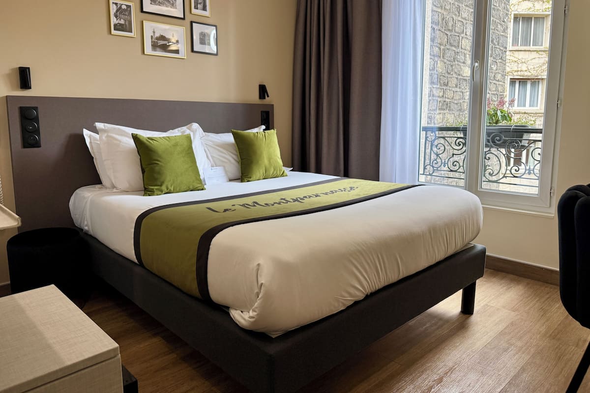 A hotel room with a bed, a bedside table, a chair, a window with a view of buildings, and framed pictures on the wall.