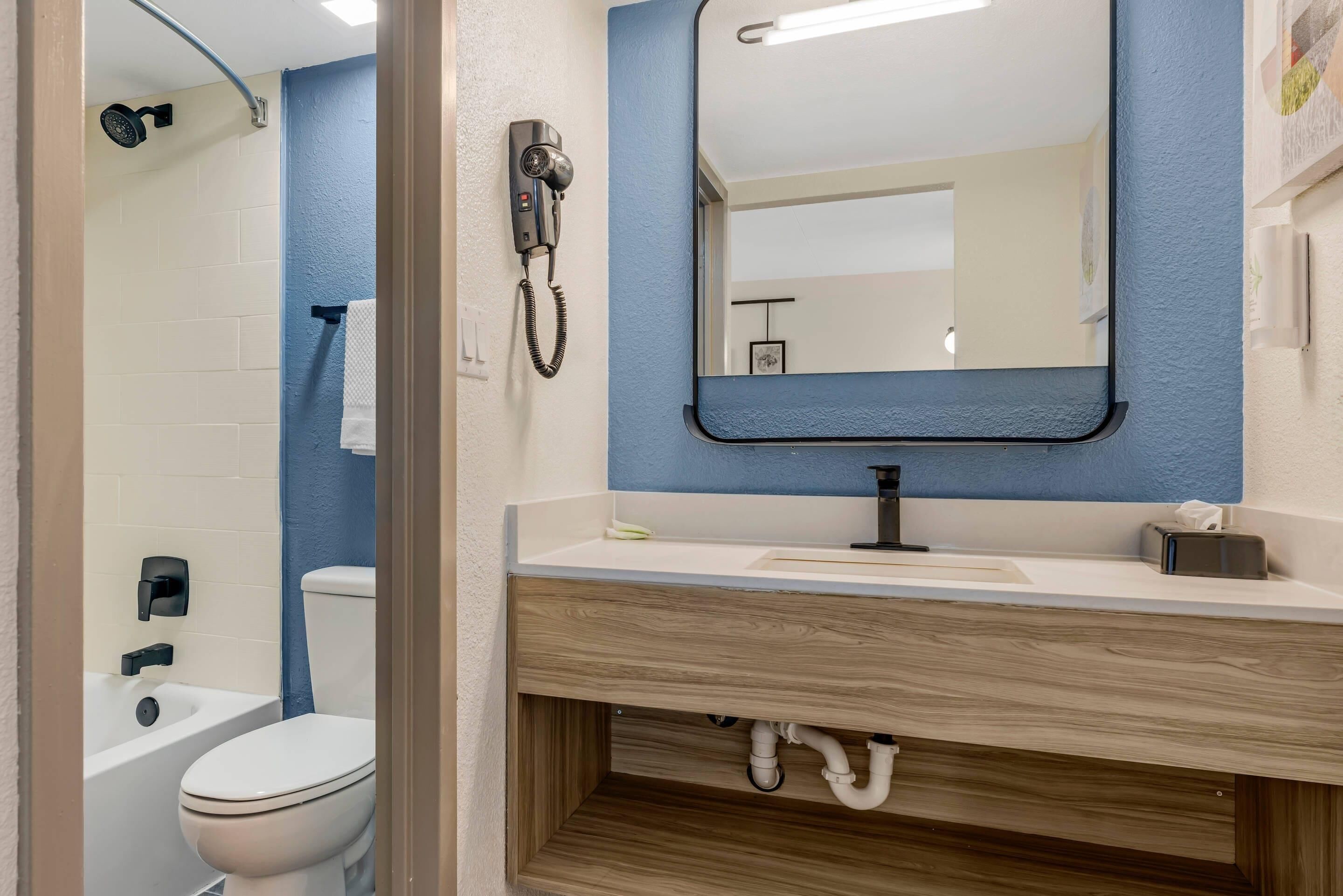 Un baño con inodoro blanco, ducha con cabezal de ducha manual, espejo con marco negro y lavamanos blanco sobre un mueble de madera.