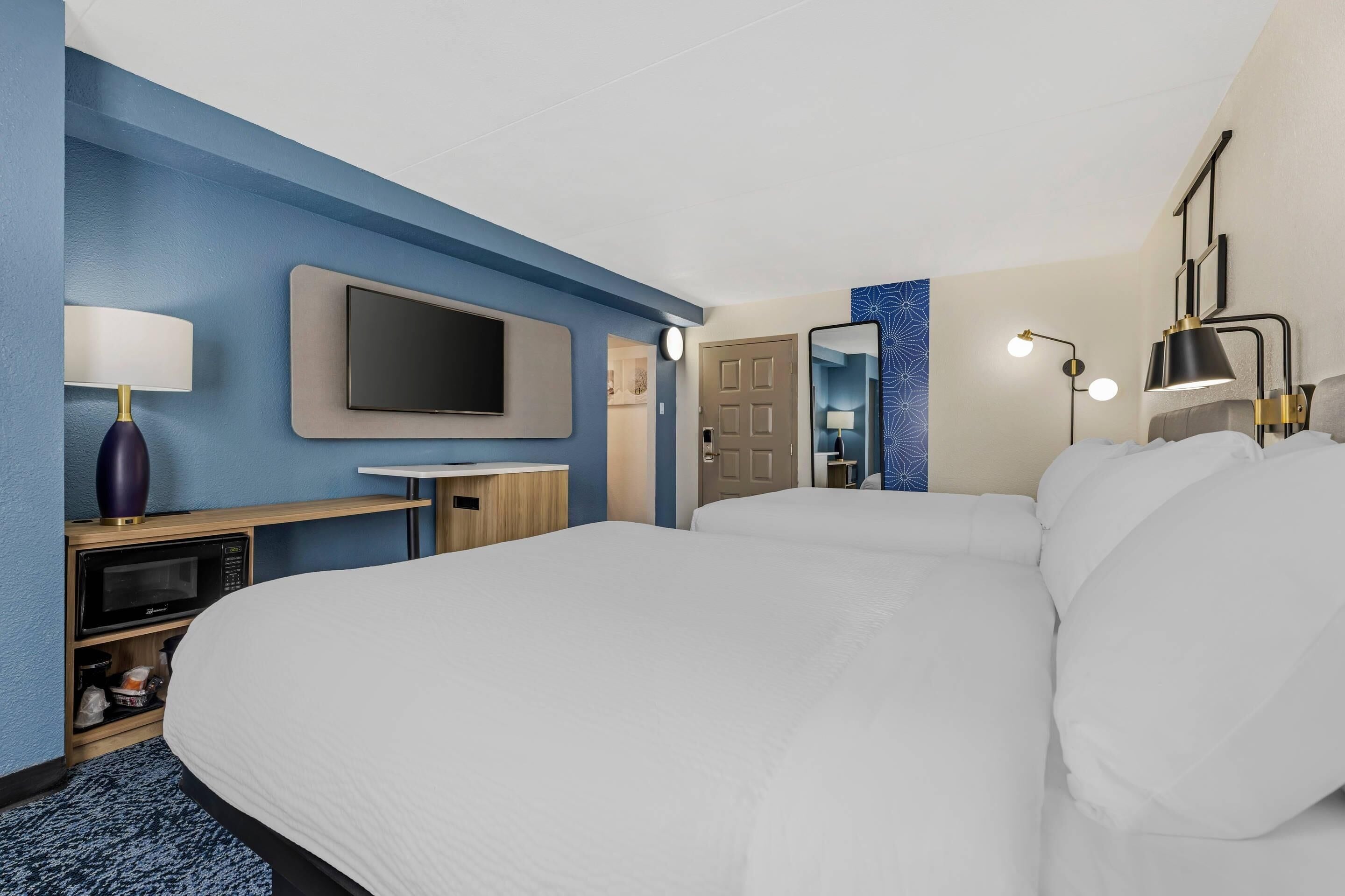 Una camera d'albergo con un letto grande, una televisione a schermo piatto, una scrivania con una lampada, un forno a microonde e una porta che conduce in un'altra stanza.