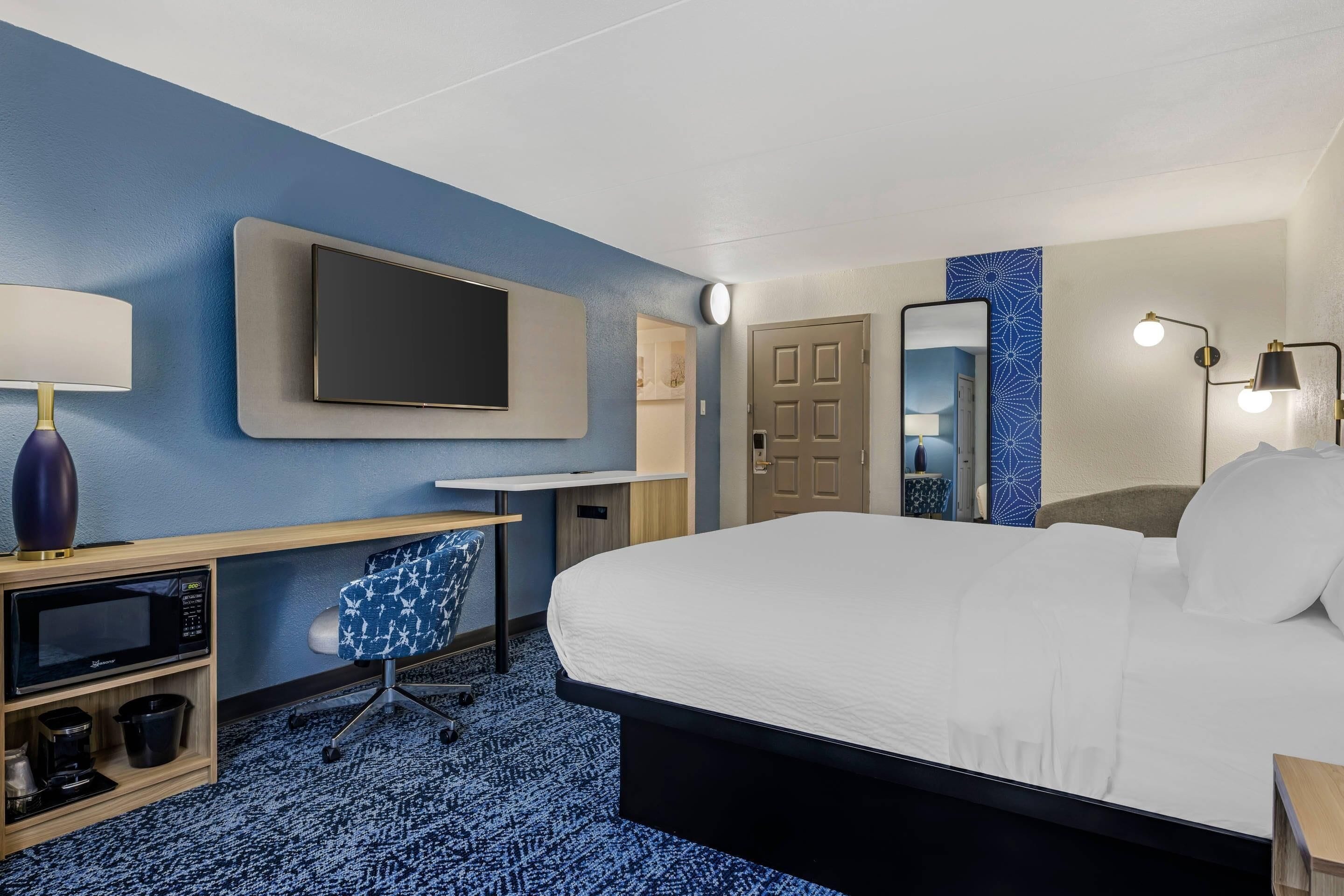 A hotel room with a bed, a desk with a flat-screen TV, a microwave, a chair, and a mirror.