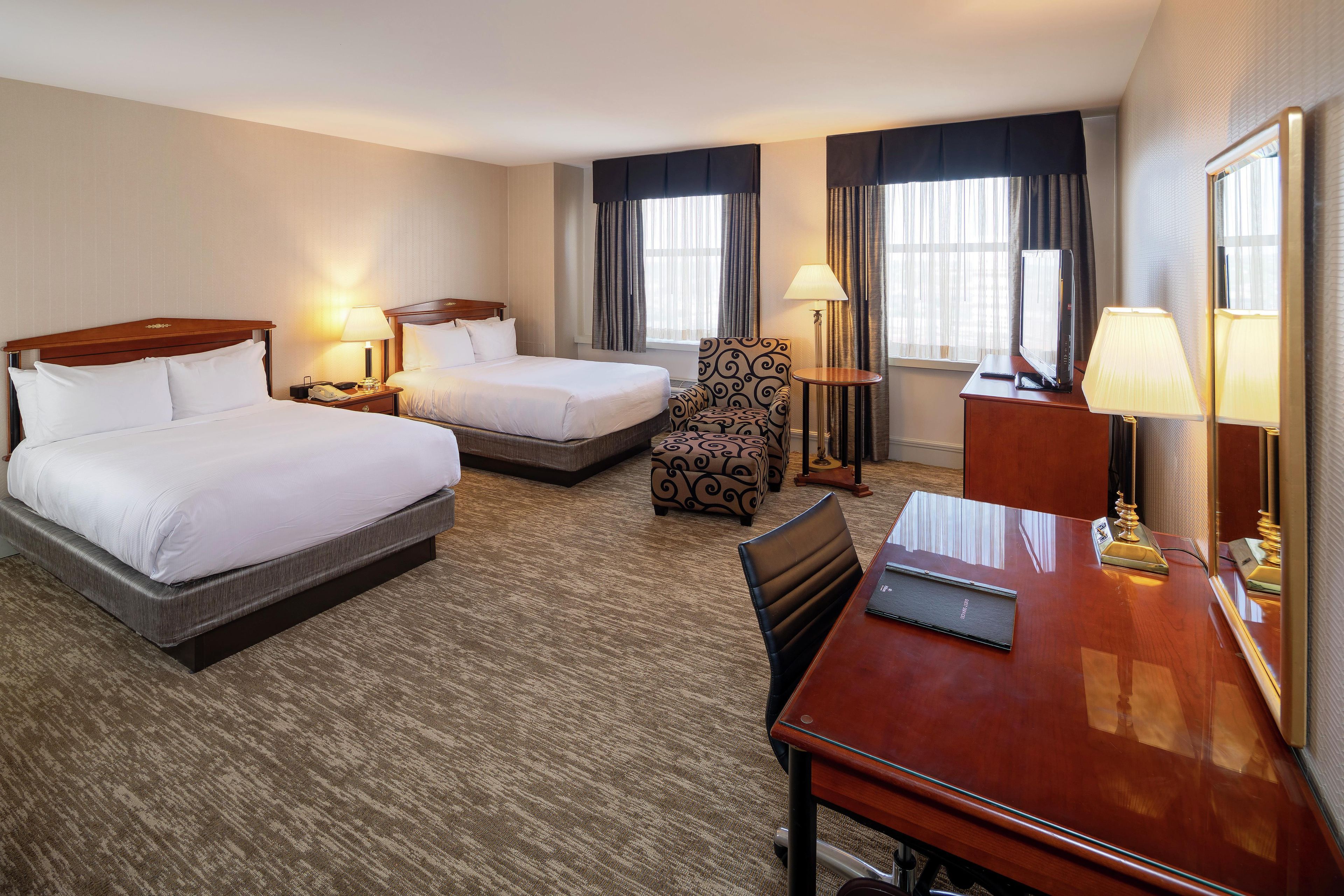 A hotel room with two beds, a desk, a chair, a television, and a lamp.