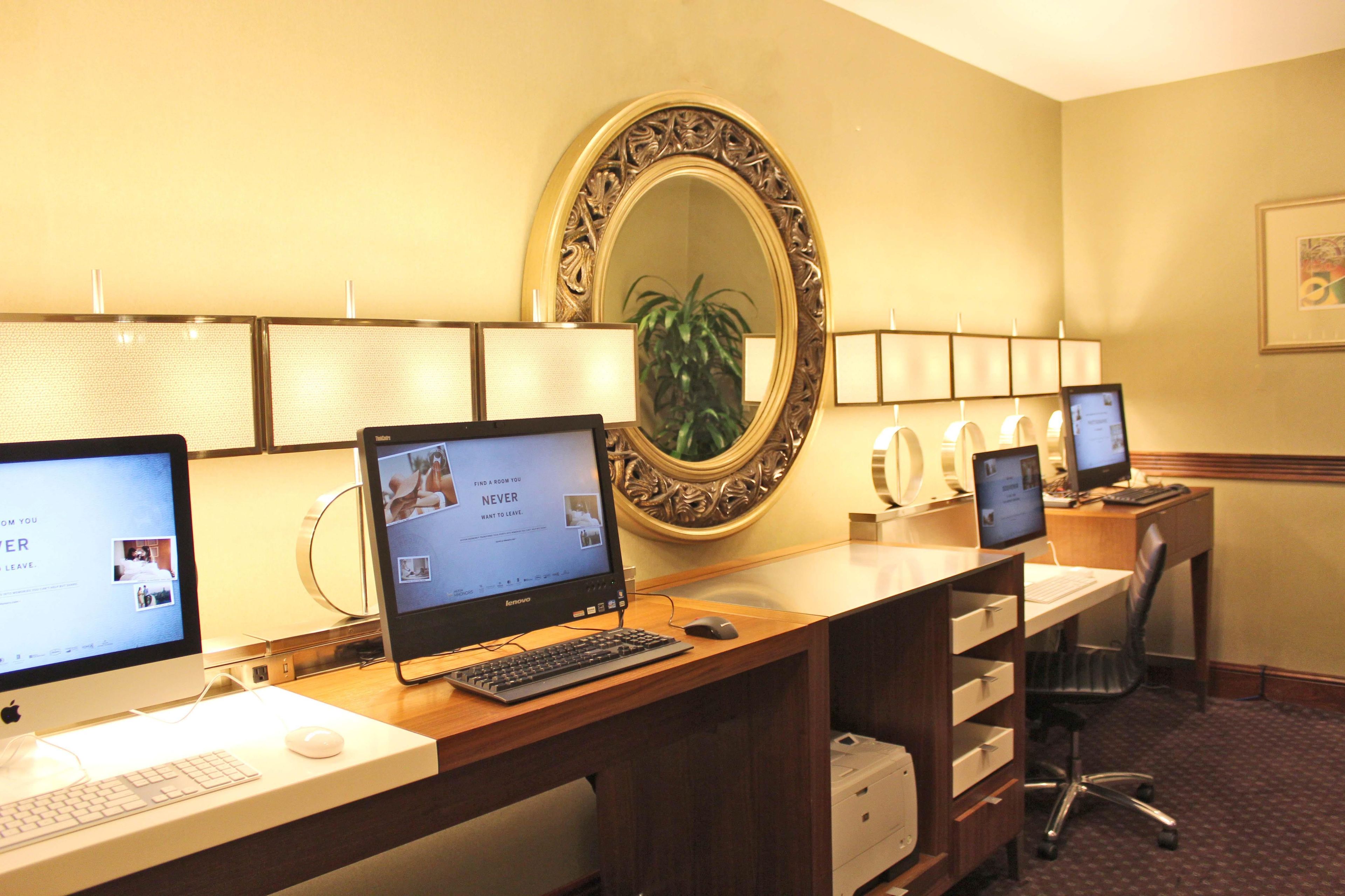 A hotel room with a desk, two monitors, a keyboard, a mouse, a printer, and a mirror.