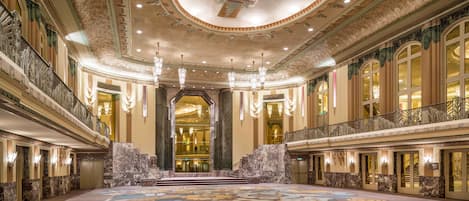 Un lobby de hotel imponente con una alfombra grande y ornamentada, techos altos con diseños intrincados y varios candelabros.