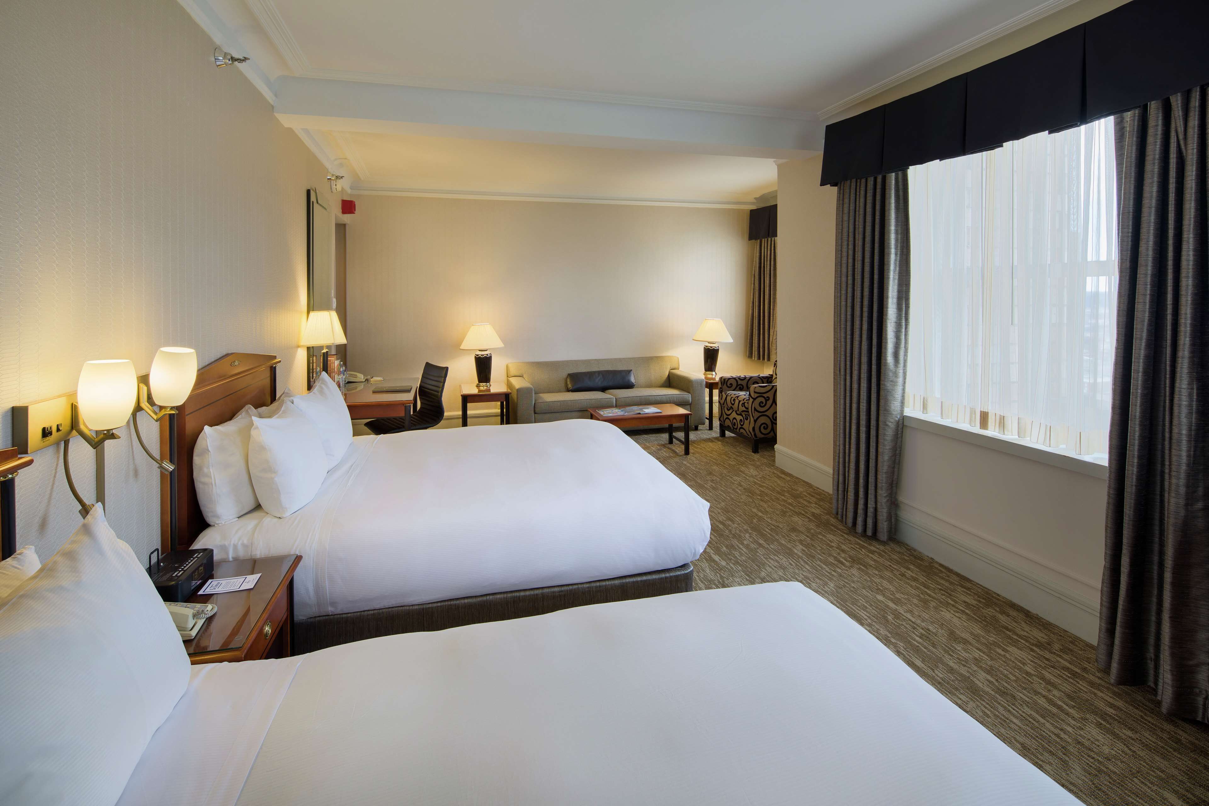 Quarto de hotel com duas camas, um sofá, uma escrivaninha e uma janela com cortinas.