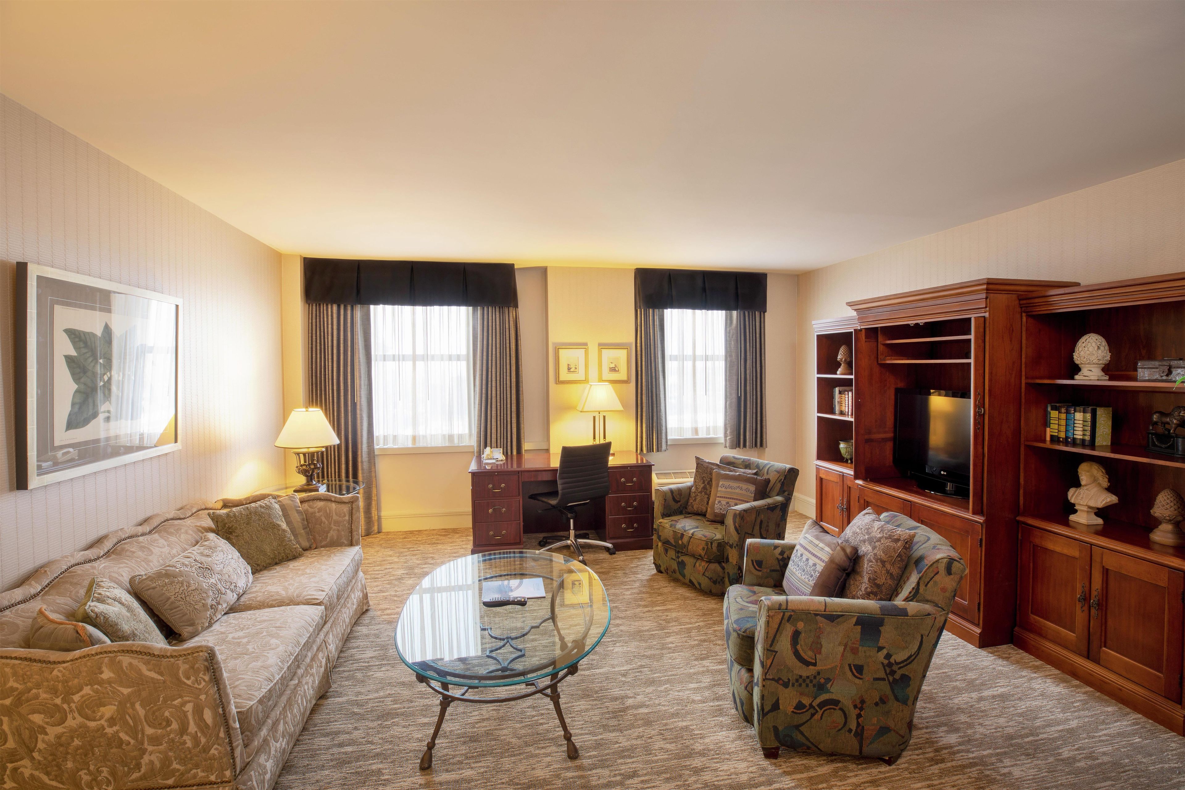 A hotel room with a sofa, armchairs, a glass coffee table, a desk with a chair, a television, and a bookshelf.