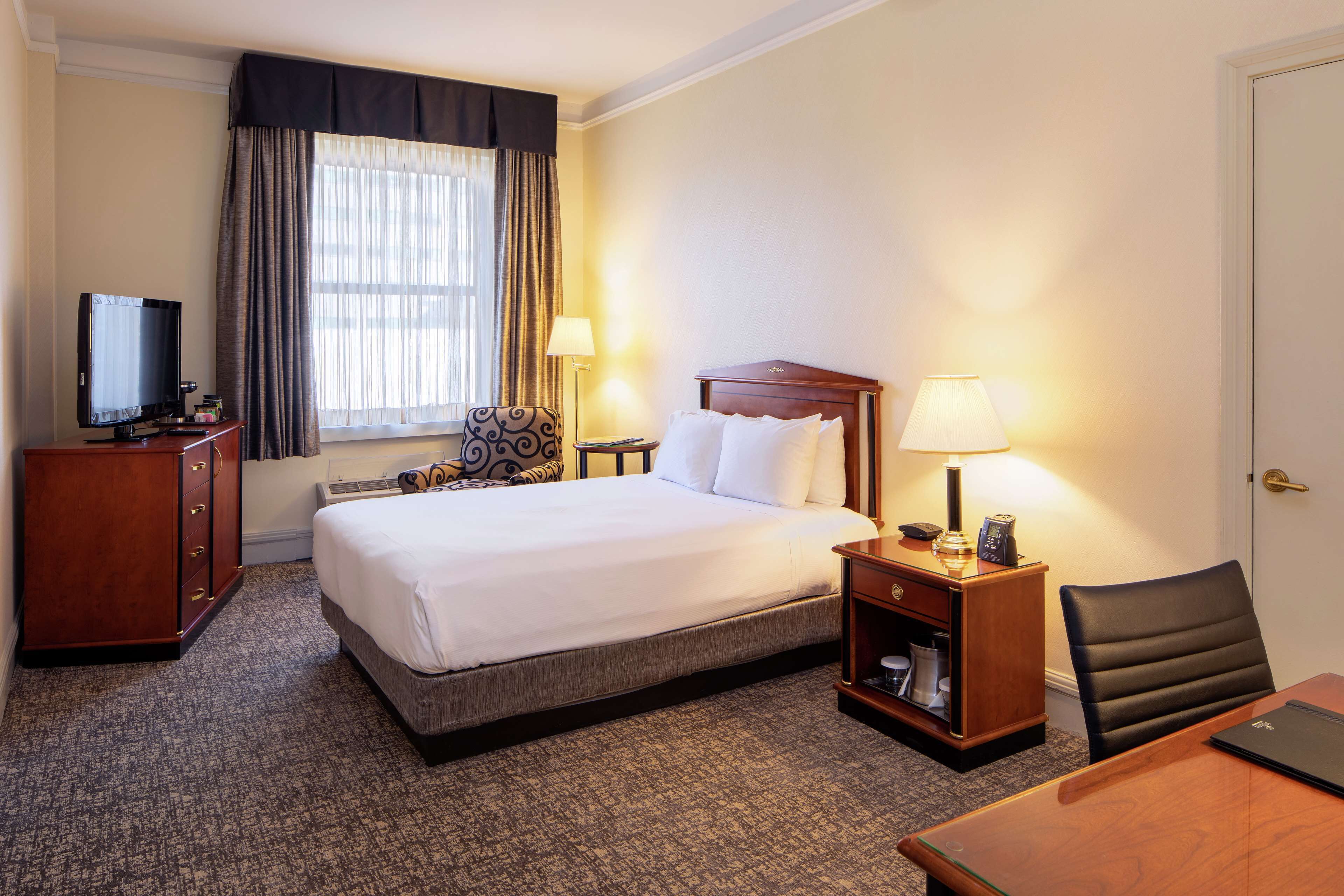 A hotel room with a bed, a desk, a chair, a television, and a lamp.