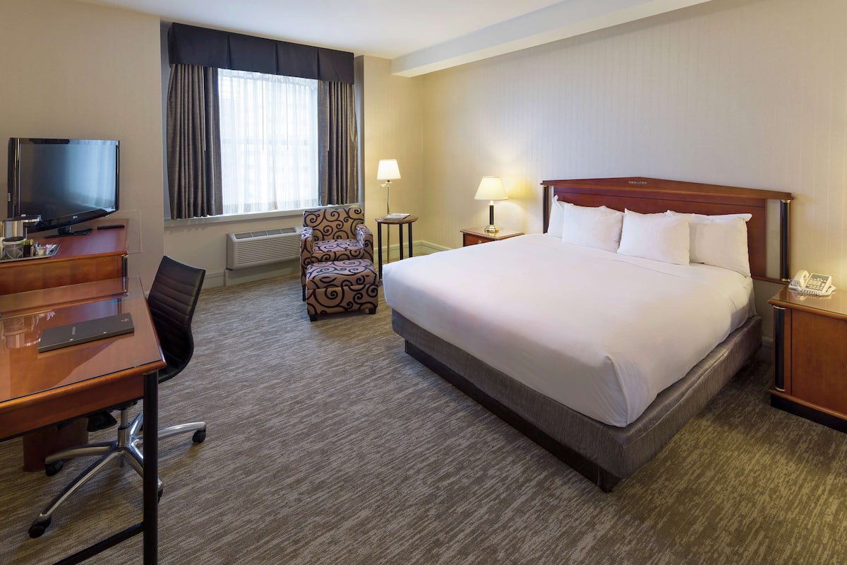 A hotel room with a large bed, a desk with a TV, a chair, a lamp, and a telephone.