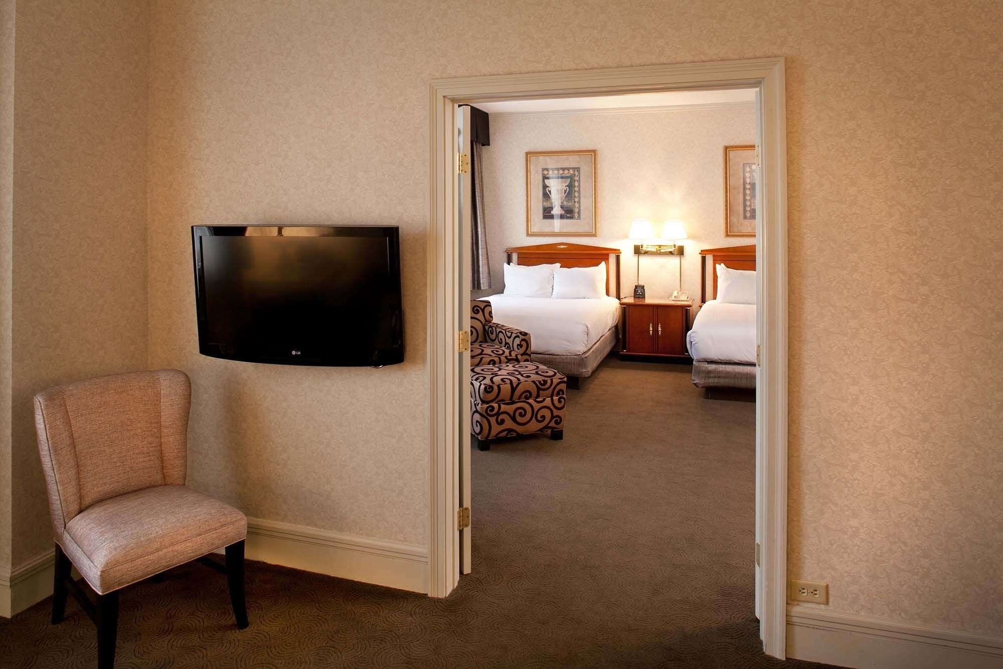 Une chambre d’hôtel dotée d’un téléviseur à écran plat fixé au mur, d’un fauteuil à motifs, d’un lit avec une literie blanche, de deux tables de chevet avec des lampes et d’un tableau encadré au mur.