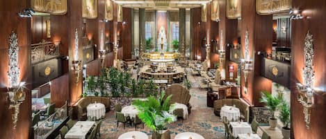 A luxurious hotel lobby with a central bar, multiple dining areas, and ornate wood paneling.