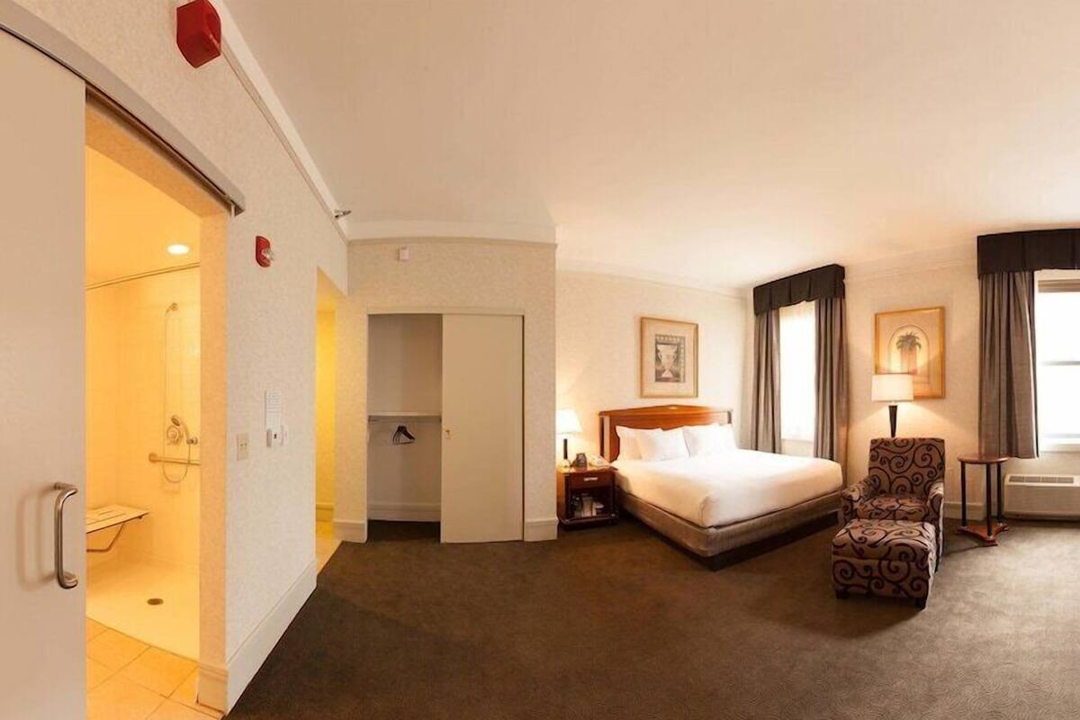 A hotel room with a bed, a chair, a nightstand, a lamp, and a window with curtains.