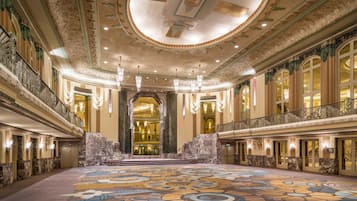 A grand hotel lobby with a large, ornate carpet, high ceilings with intricate designs, and a central staircase leading to an upper level.