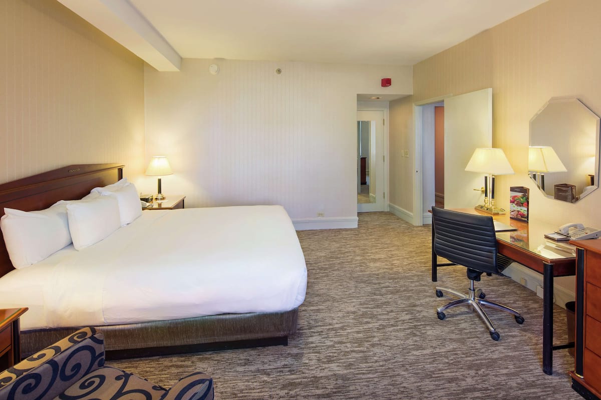 A hotel room with a large bed, a desk with a chair, a mirror, and a lamp.