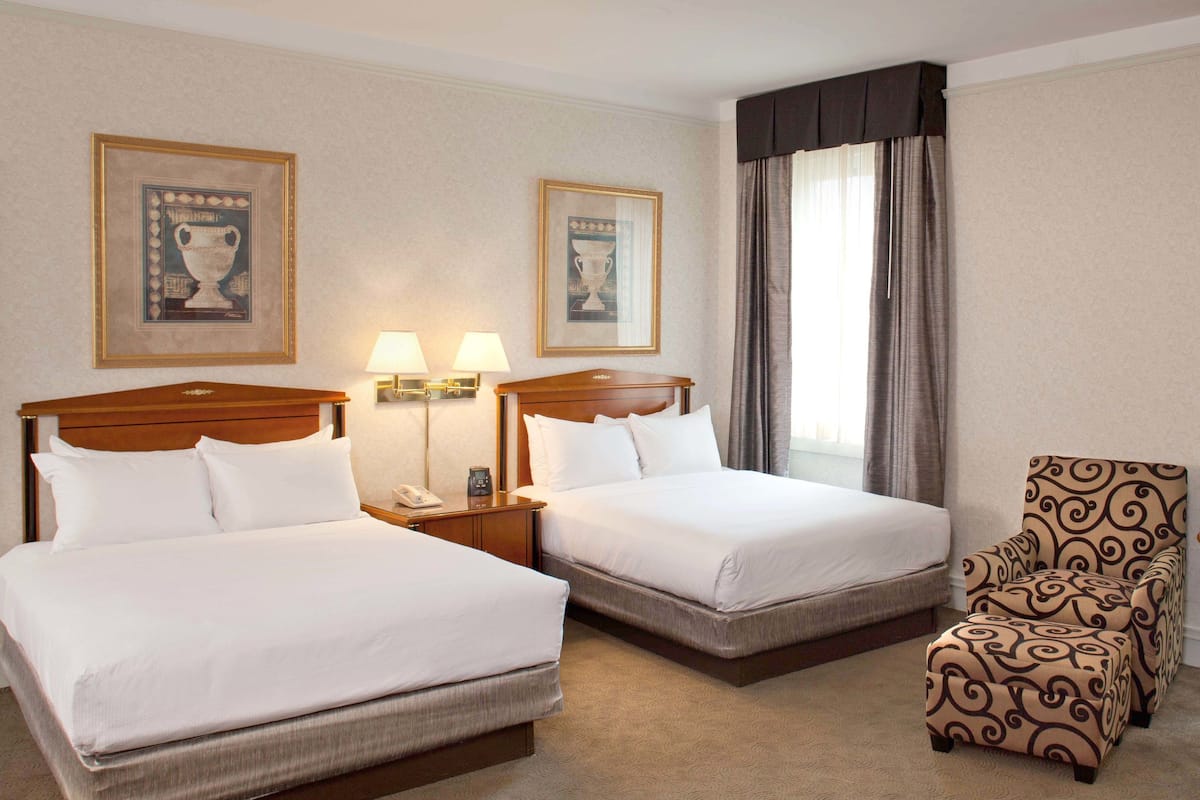 A hotel room with two beds, a chair, and two lamps.