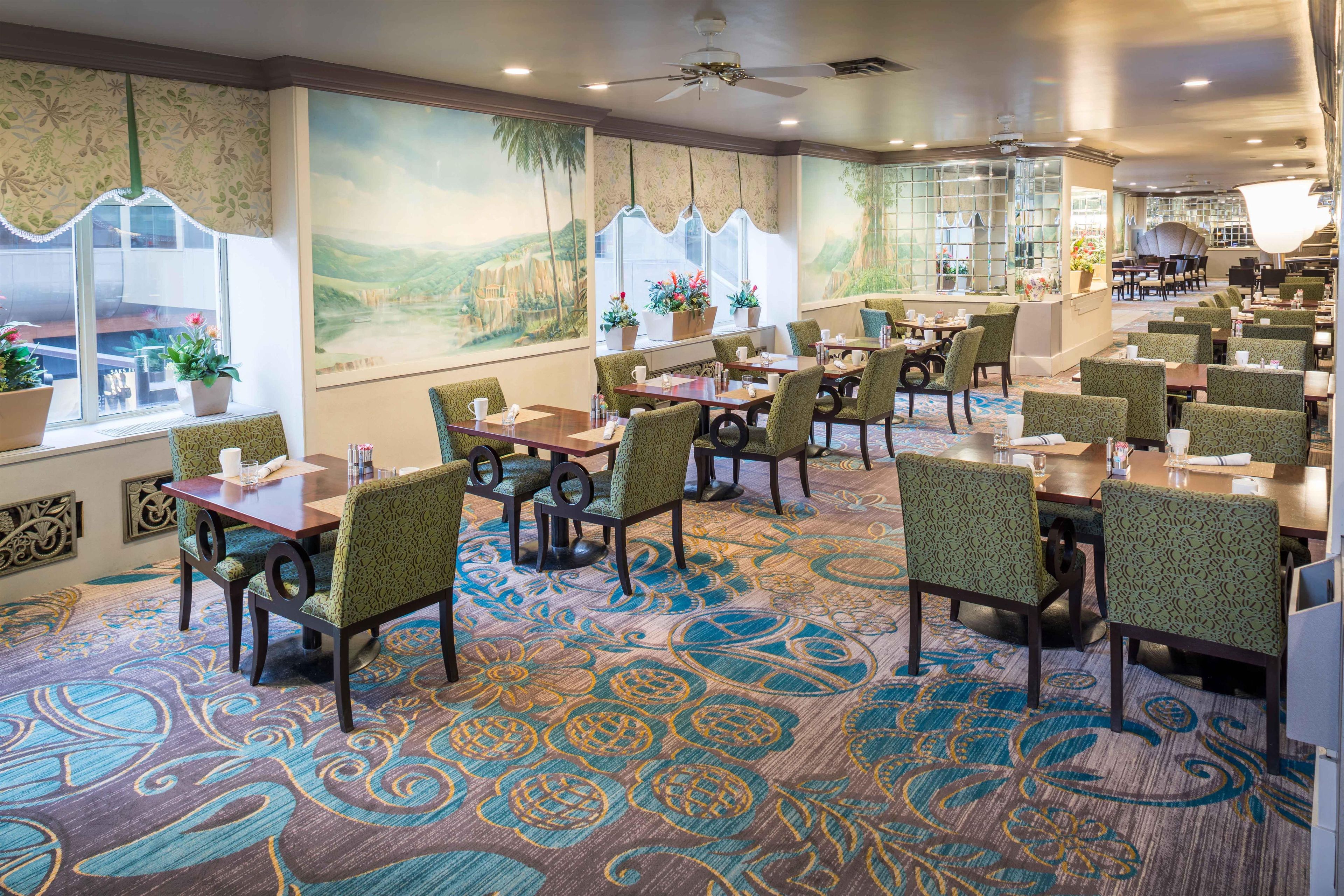 A restaurant with patterned carpet, green upholstered chairs, and a mural of a landscape.