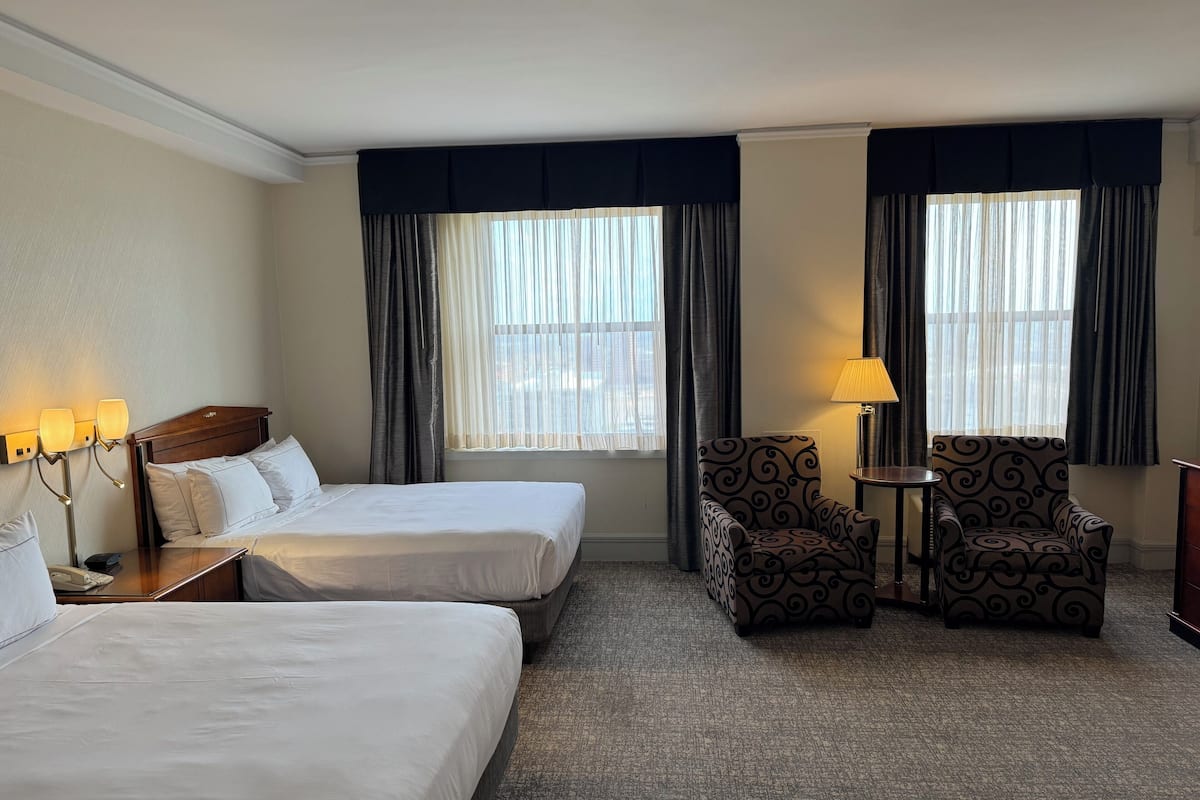 A hotel room with two beds, a chair, a table, and a lamp.