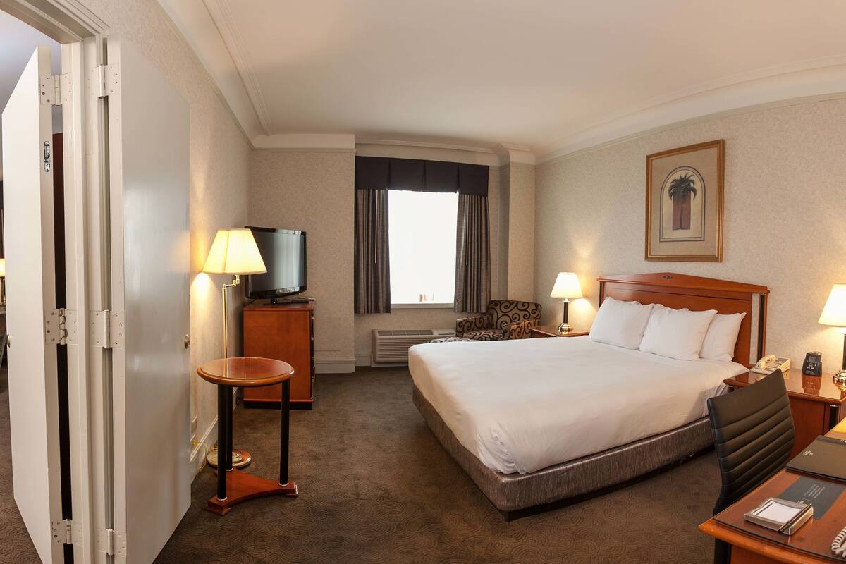 A hotel room with a large bed, two bedside tables with lamps, a TV, a desk with a chair, and a window with curtains.