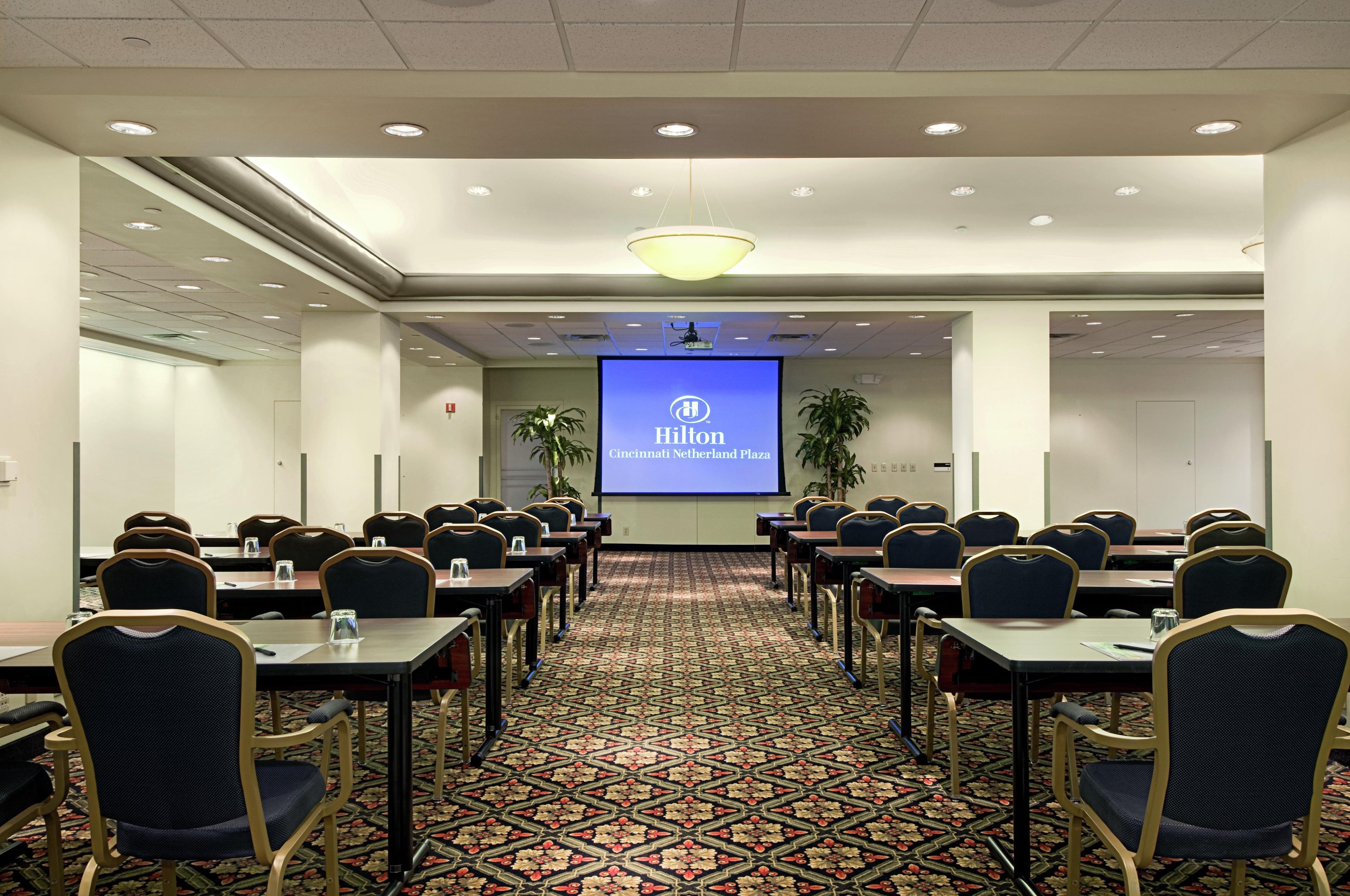 Un salón de conferencias con una pantalla de proyector mostrando el logotipo y la ubicación de Hilton, dispuesto con mesas y sillas para reuniones.