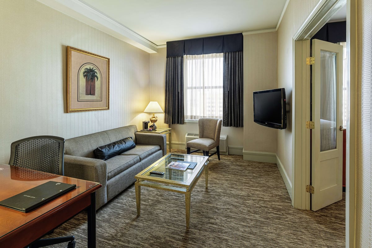 A hotel room with a sofa, armchair, glass coffee table, TV, and a door with a window.