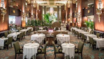 Une salle à manger spacieuse et élégamment décorée, avec plusieurs tables et chaises rondes, agrémentée d’une fontaine décorative centrale et d’une végétation luxuriante.