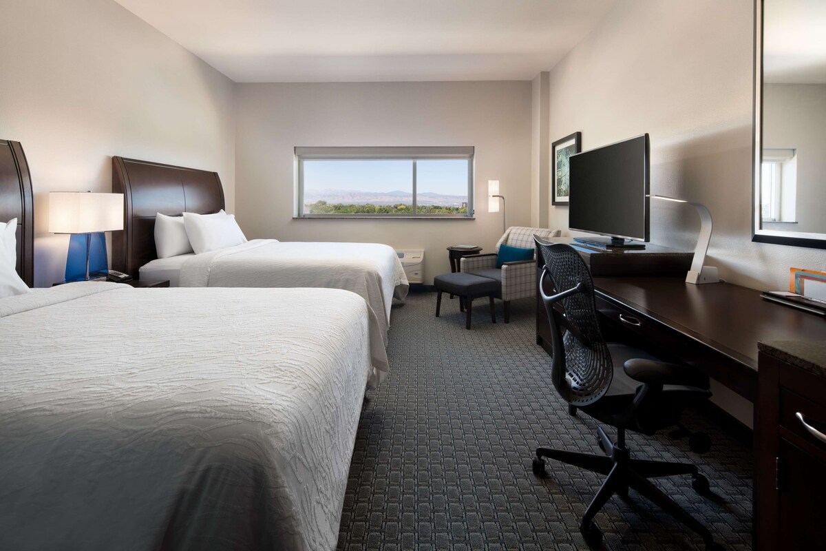 A hotel room with two beds, a desk with a computer, a TV, a chair, and a window with a view.