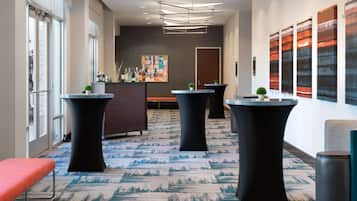 A modern hotel lobby with a reception desk, three black round tables with potted plants, a red bench, and a large abstract painting on the wall.