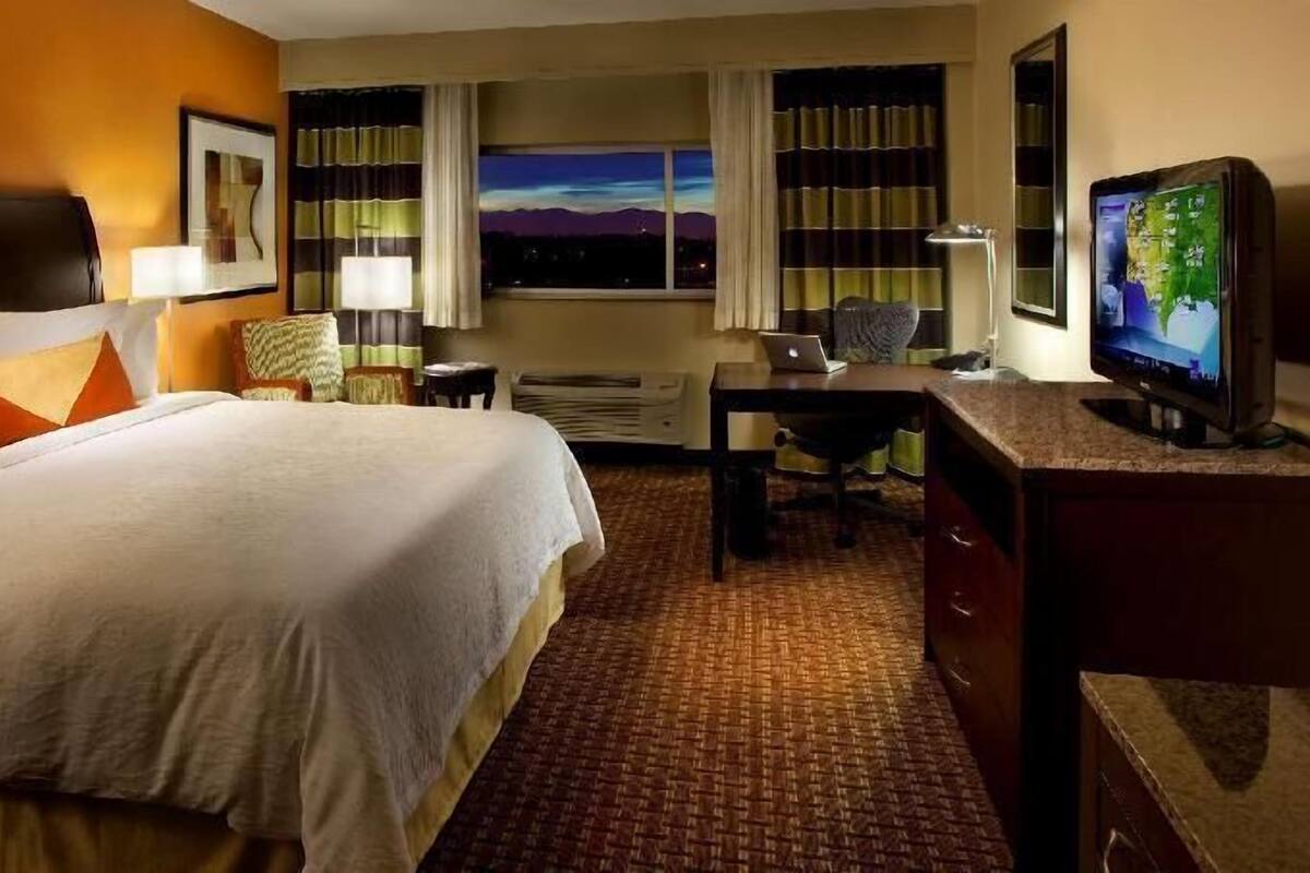 A hotel room with a bed, desk, chair, TV, and a window with a cityscape view.