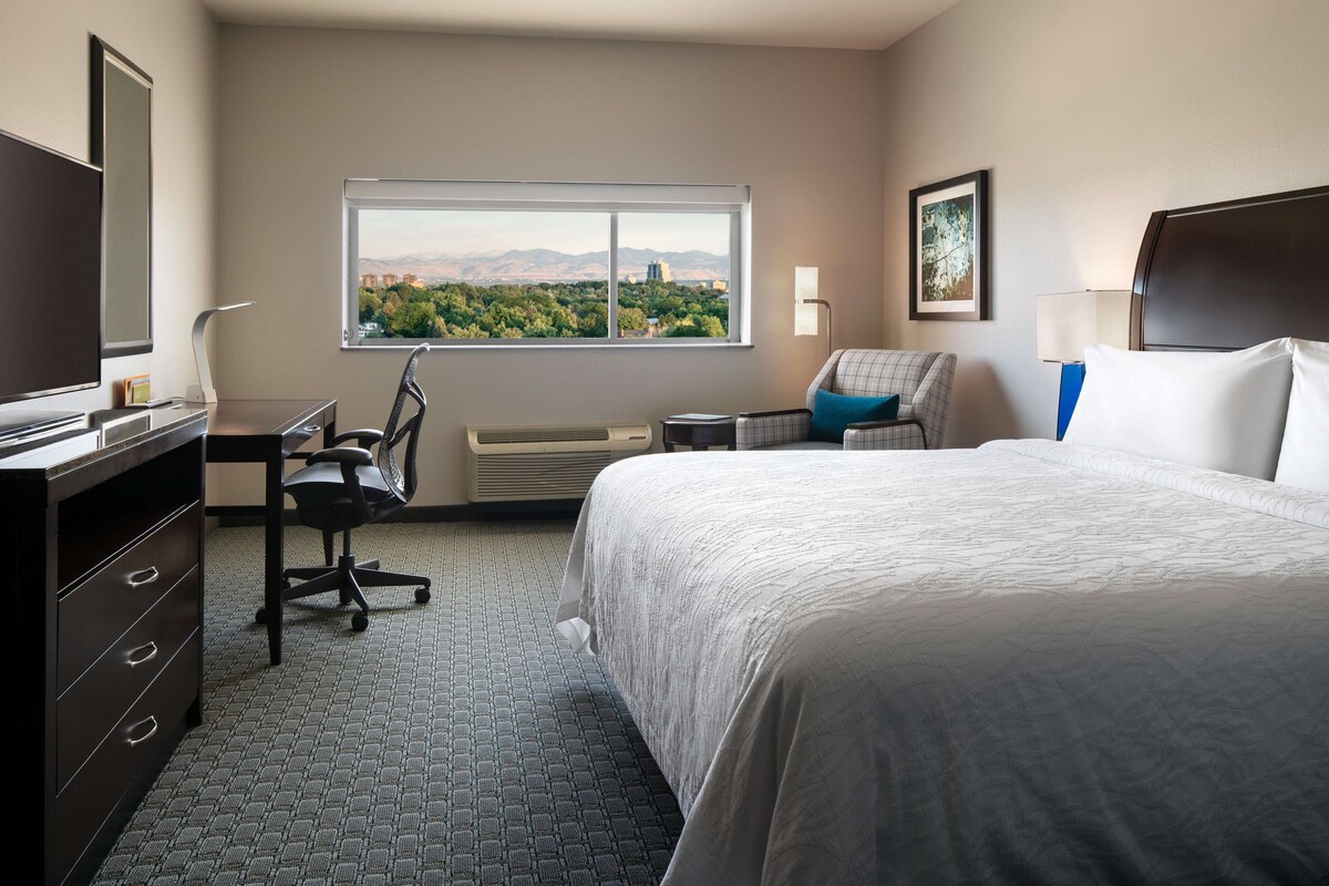 A hotel room with a large bed, a desk with a chair, a window with a city view, and a flat-screen TV.