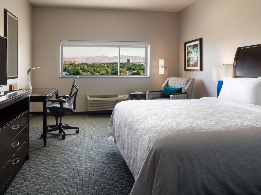 A hotel room with a large bed, a desk with a chair, a window with a city view, and a flat-screen TV.