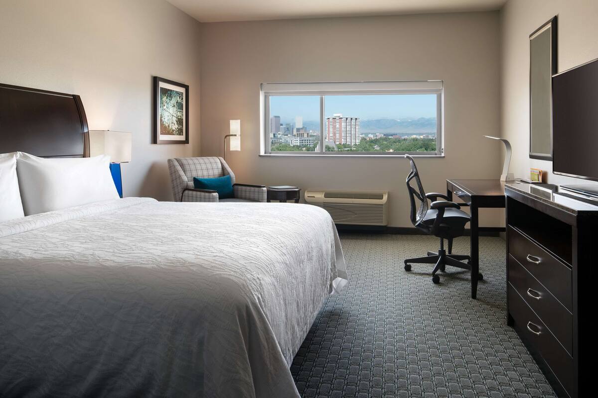 A hotel room with a large bed, a desk, a chair, a TV, and a window with a city view.