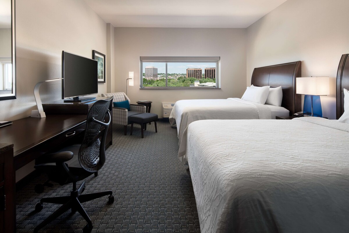 A hotel room with a desk, two beds, a chair, a TV, and a window with a city view.