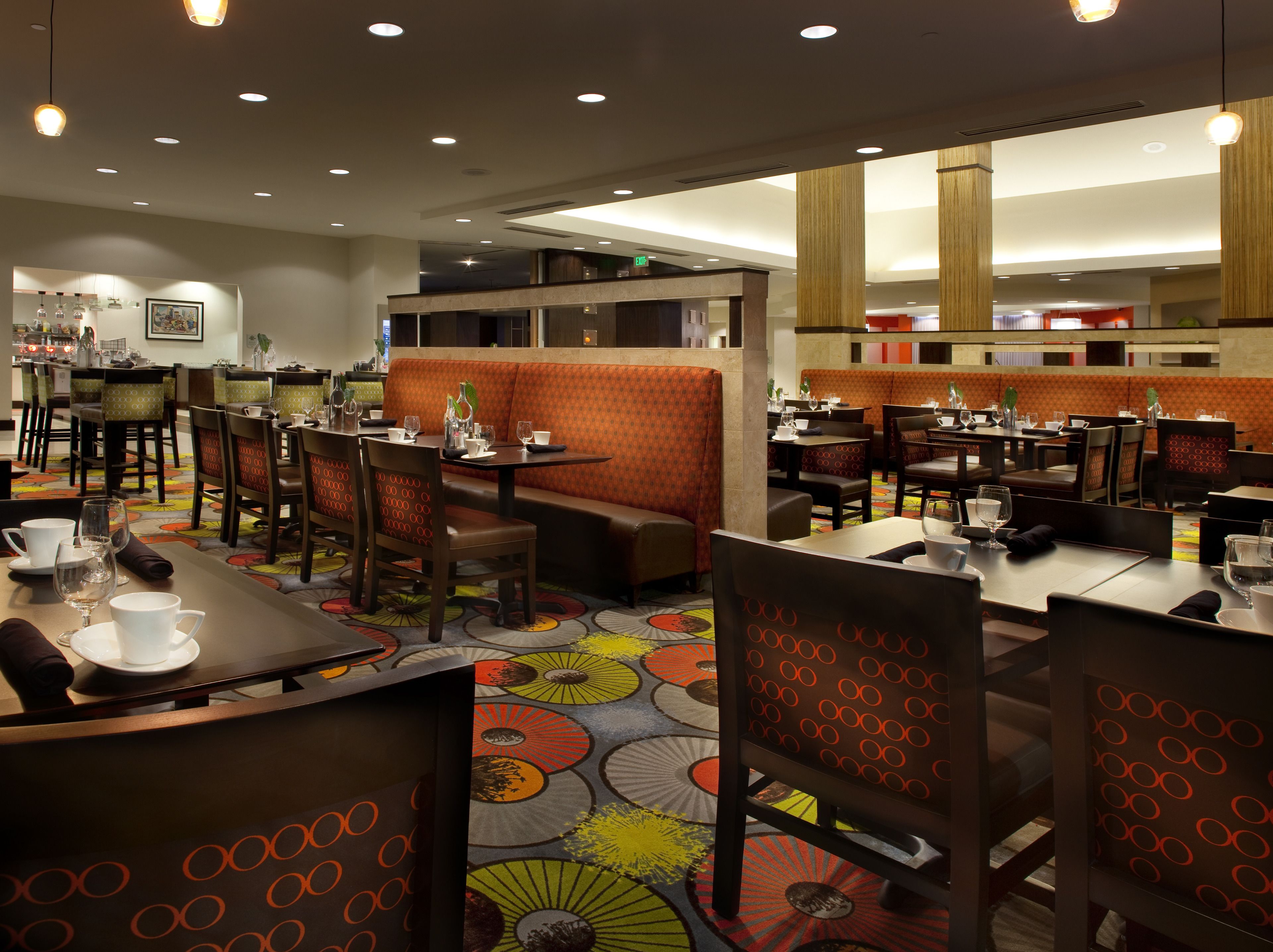 A restaurant with booths and tables, some with orange cushions. There are chairs with a circular pattern, and the floor has a colorful design.
