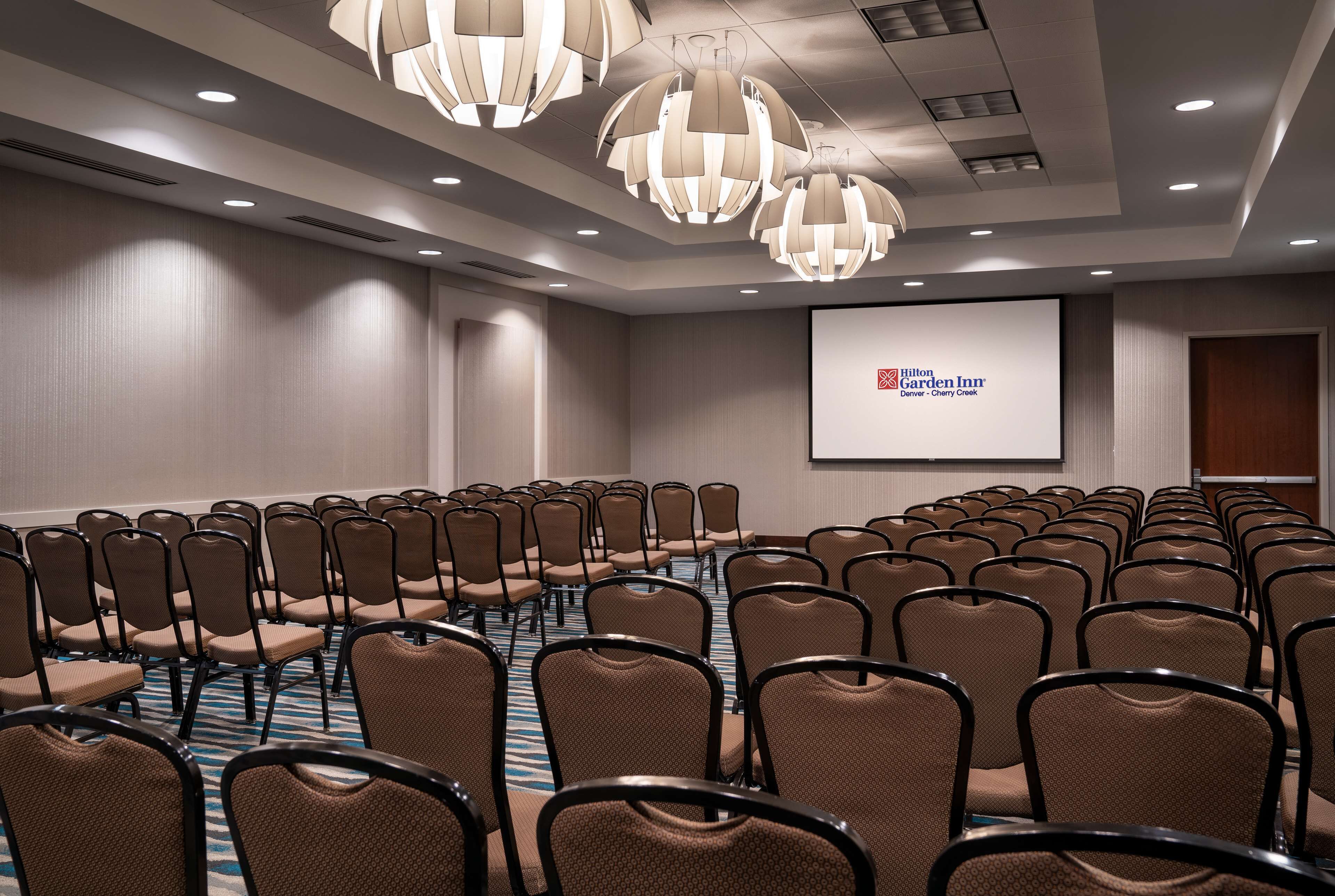 A conference room with rows of chairs, a projector screen displaying 'Hilton Garden Inn Denver - Cherry Creek', and modern pendant lighting.