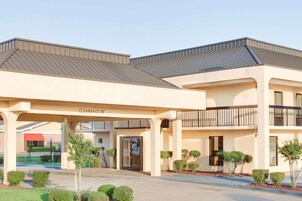 Entrada de hotel com uma galeria coberta, uma placa indicando 'CLEARANCE 96', e um estacionamento com algumas árvores e arbustos.