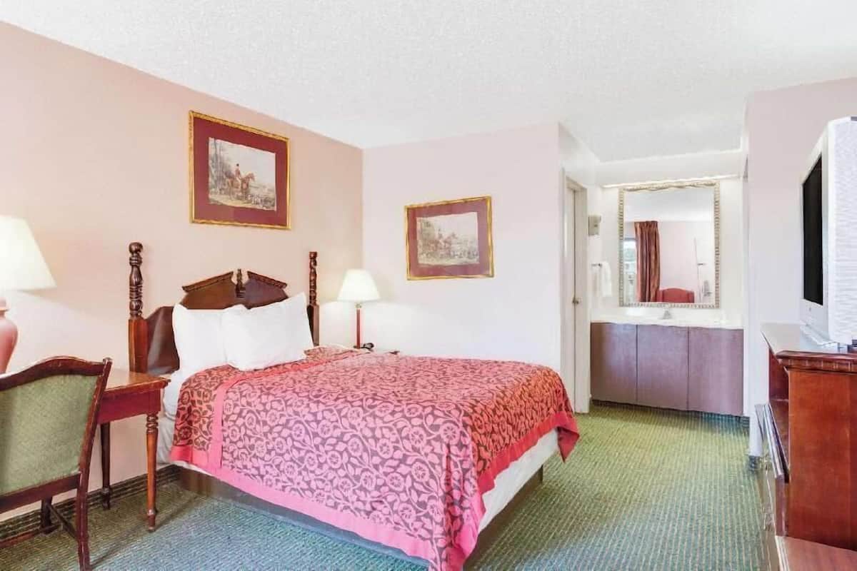 A hotel room with a bed, a chair, a desk, a mirror, and two framed pictures on the wall.