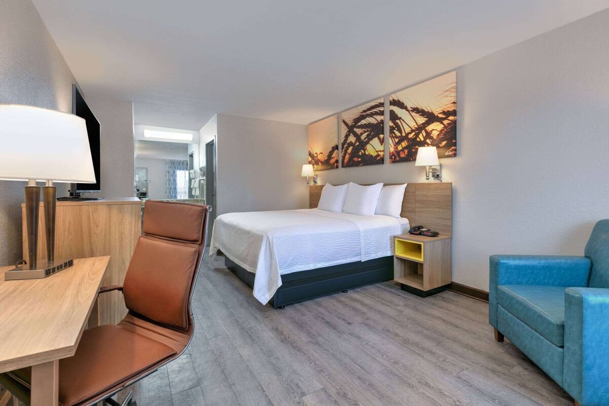 A modern hotel room with a bed, a desk, a chair, a nightstand, and a wall-mounted lamp.