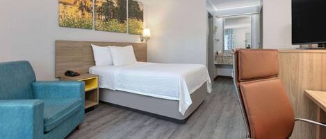 A hotel room with a bed, a blue armchair, a brown chair, a TV, and artwork on the wall.