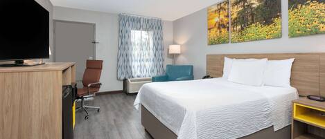 A hotel room with a bed, a desk with a TV, a chair, a window with curtains, and wall art.