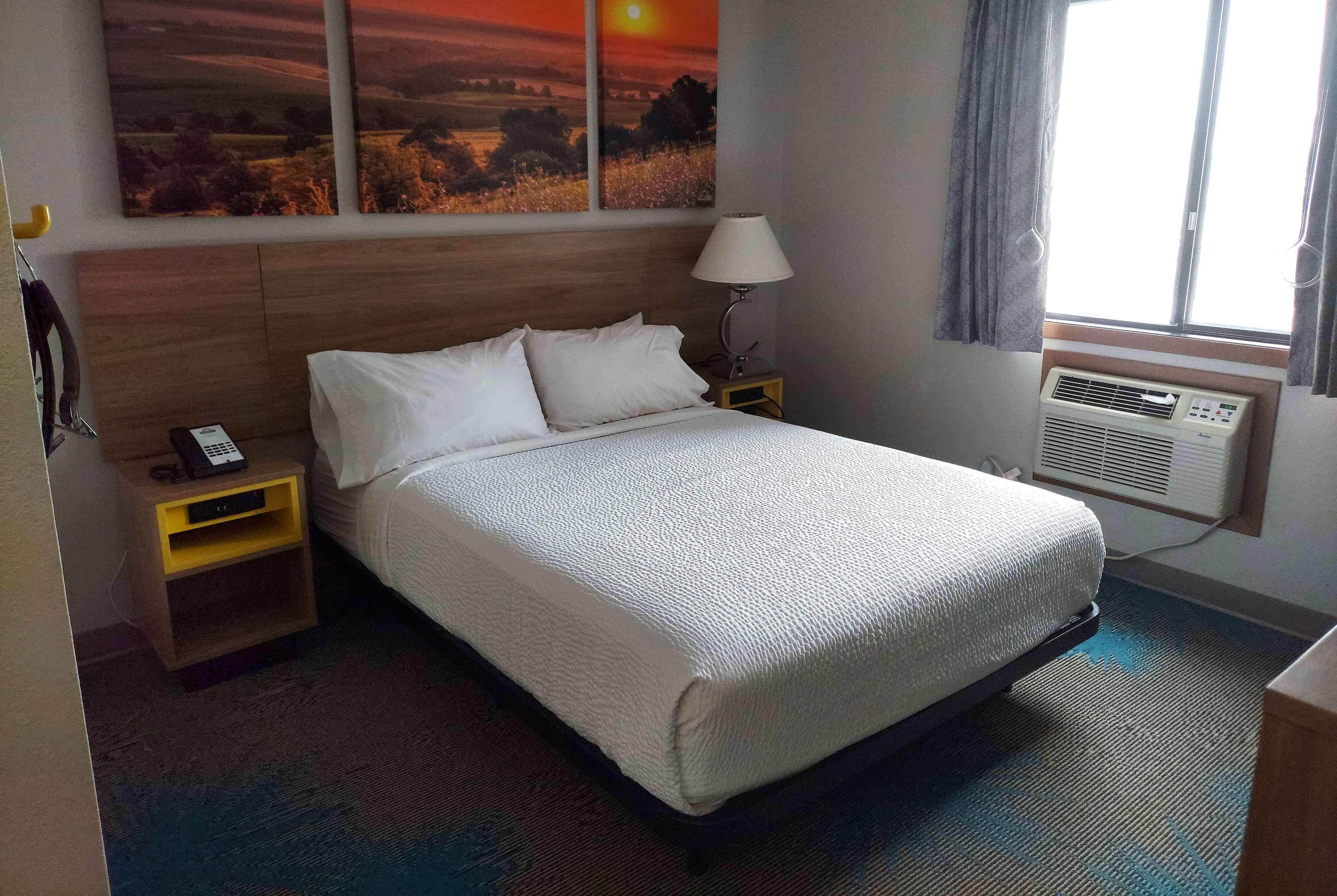 Habitació d'hotel amb un llit, una taula de nit amb una làmpada, aire condicionat fixat a la paret i una finestra amb cortines.