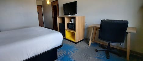 A hotel room with a bed, a desk with a chair, a television, and a small shelf.