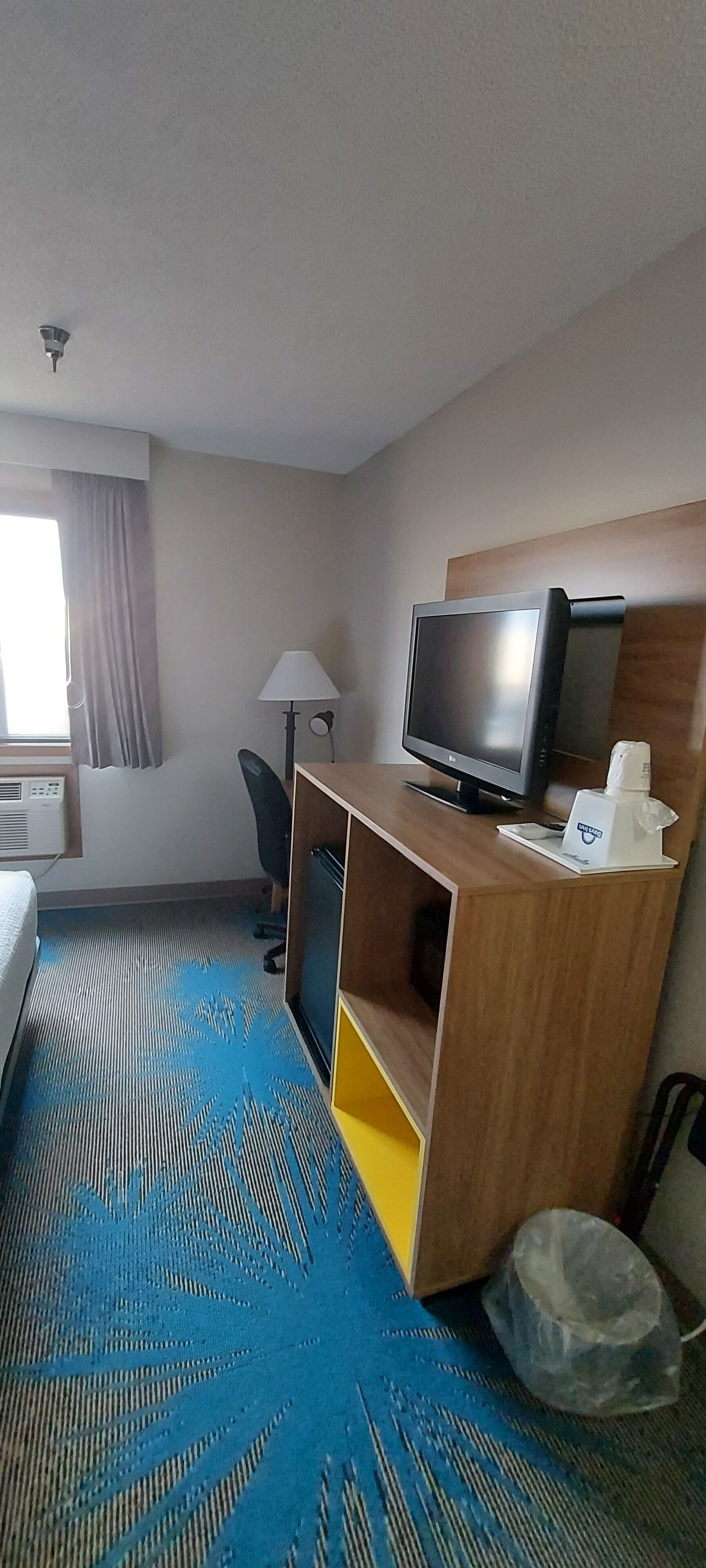 Ein Hotelzimmer mit Bett, Schreibtisch mit Bildschirm, Stuhl, Lampe und einem Fenster mit Vorhängen.