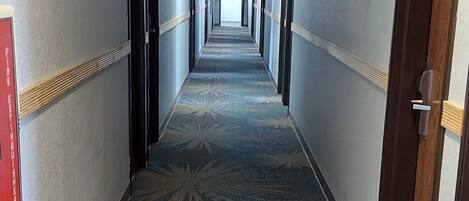A long, narrow hotel corridor with patterned carpet, doors on both sides, and a ceiling light fixture.