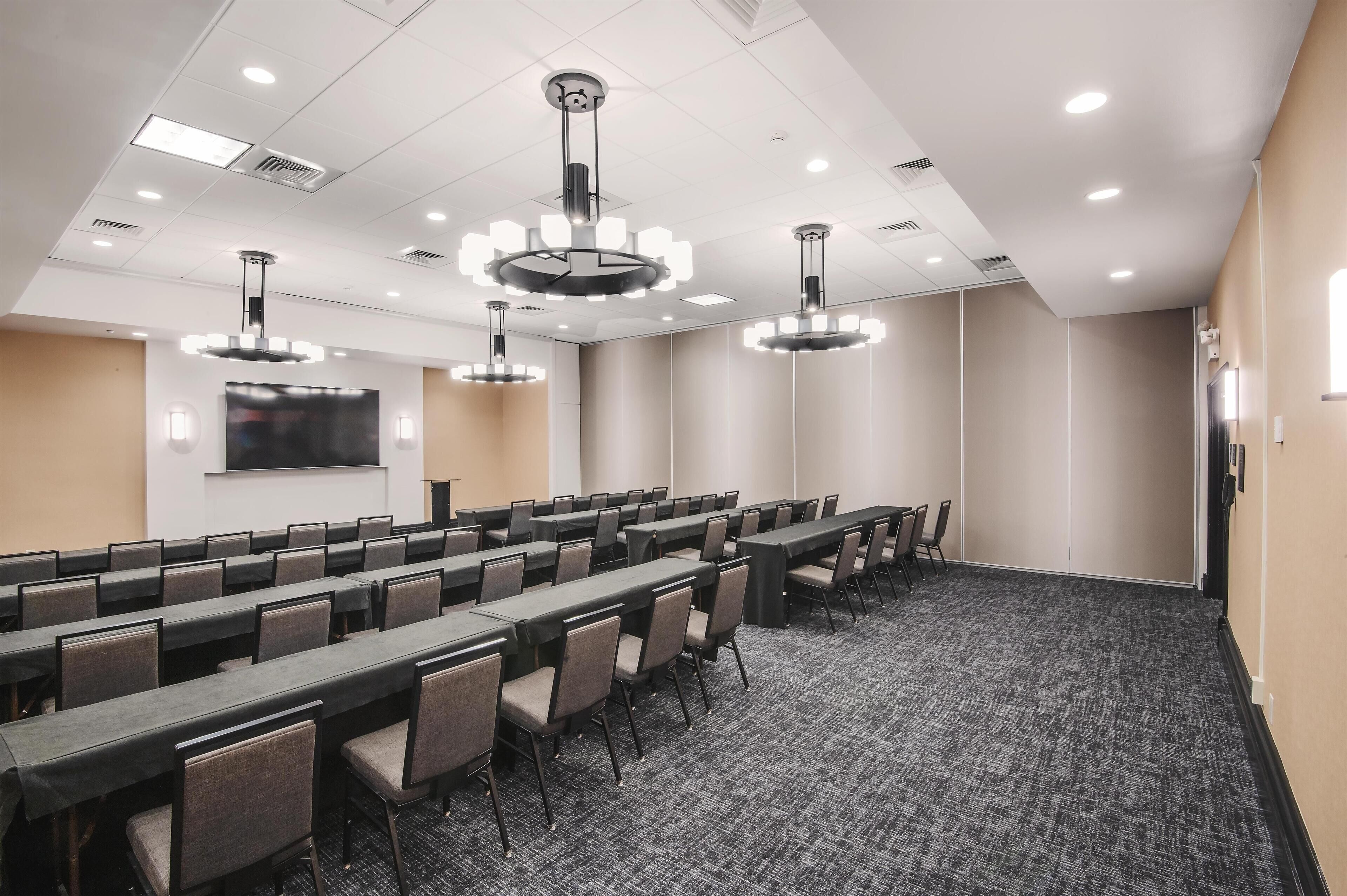 A conference room with long tables and chairs, recessed ceiling lights, and a flat-screen TV mounted on the wall.