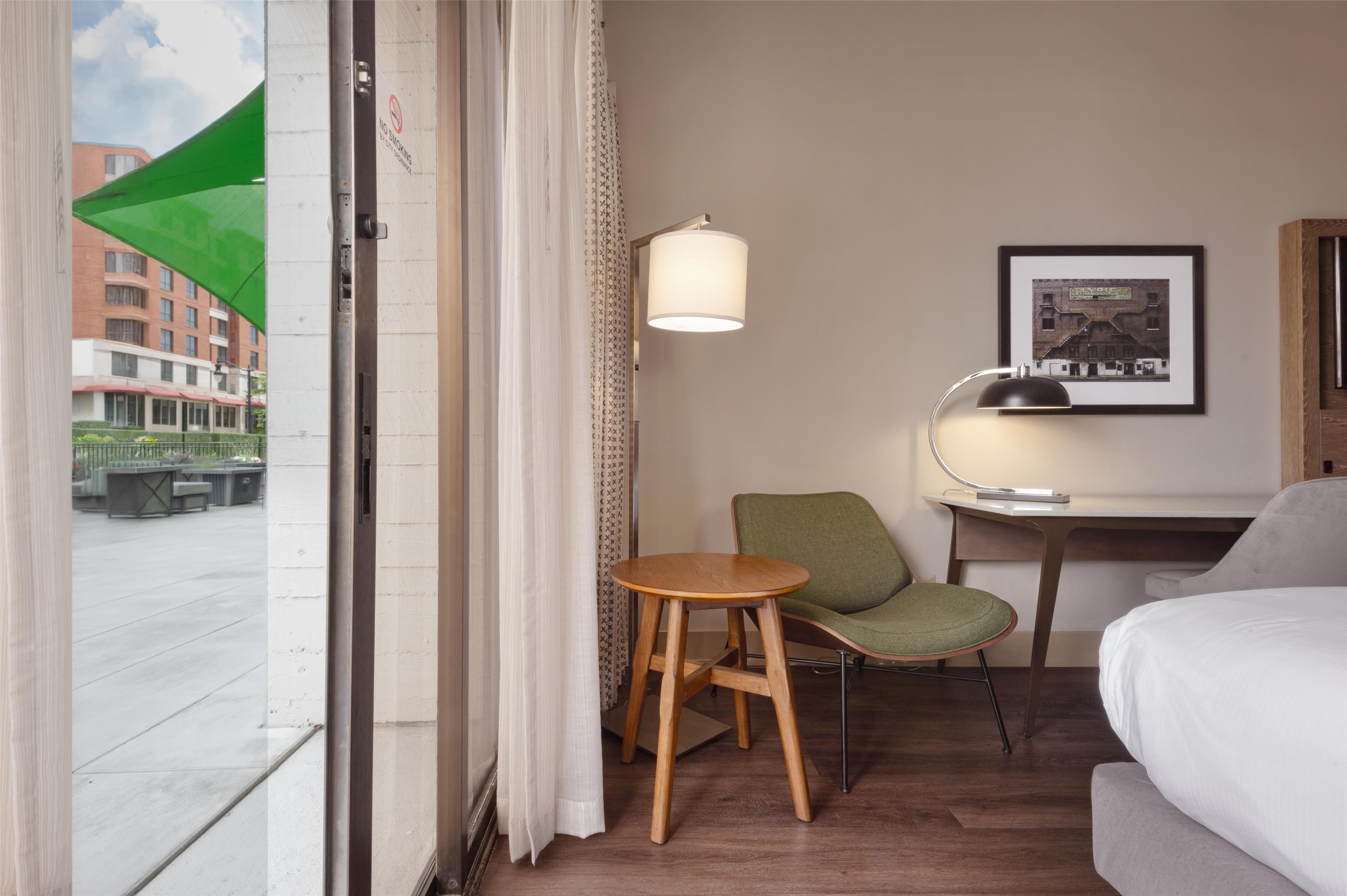 Ein Hotelzimmer mit einem Bett, einem Schreibtisch mit Lampe, einem Stuhl, einem kleinen hölzernen Hocker, einem Fenster mit Blick auf Gebäude und einem gerahmten Bild an der Wand.