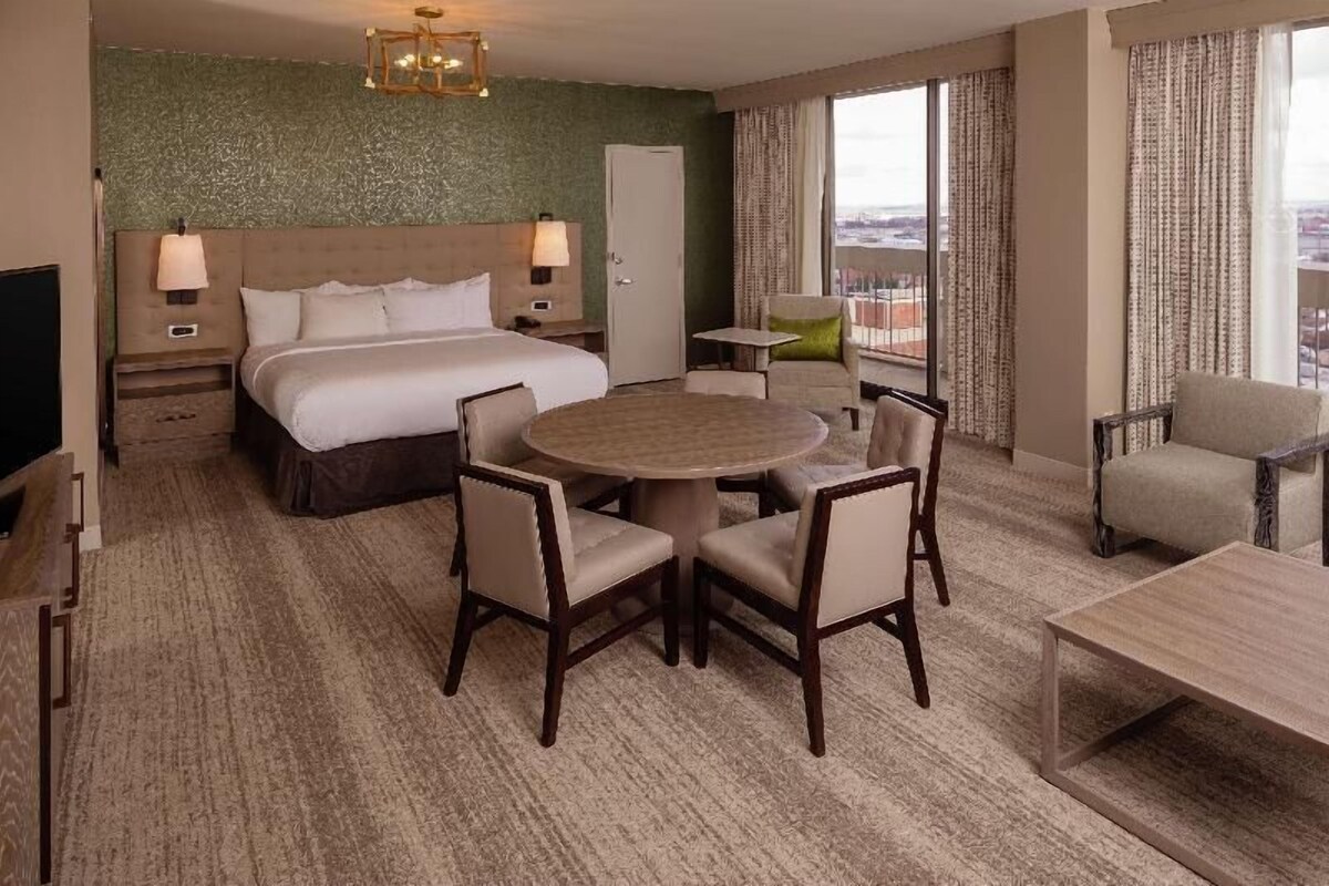 A hotel room with a bed, a round table with chairs, a TV, a desk, and a view of the city.