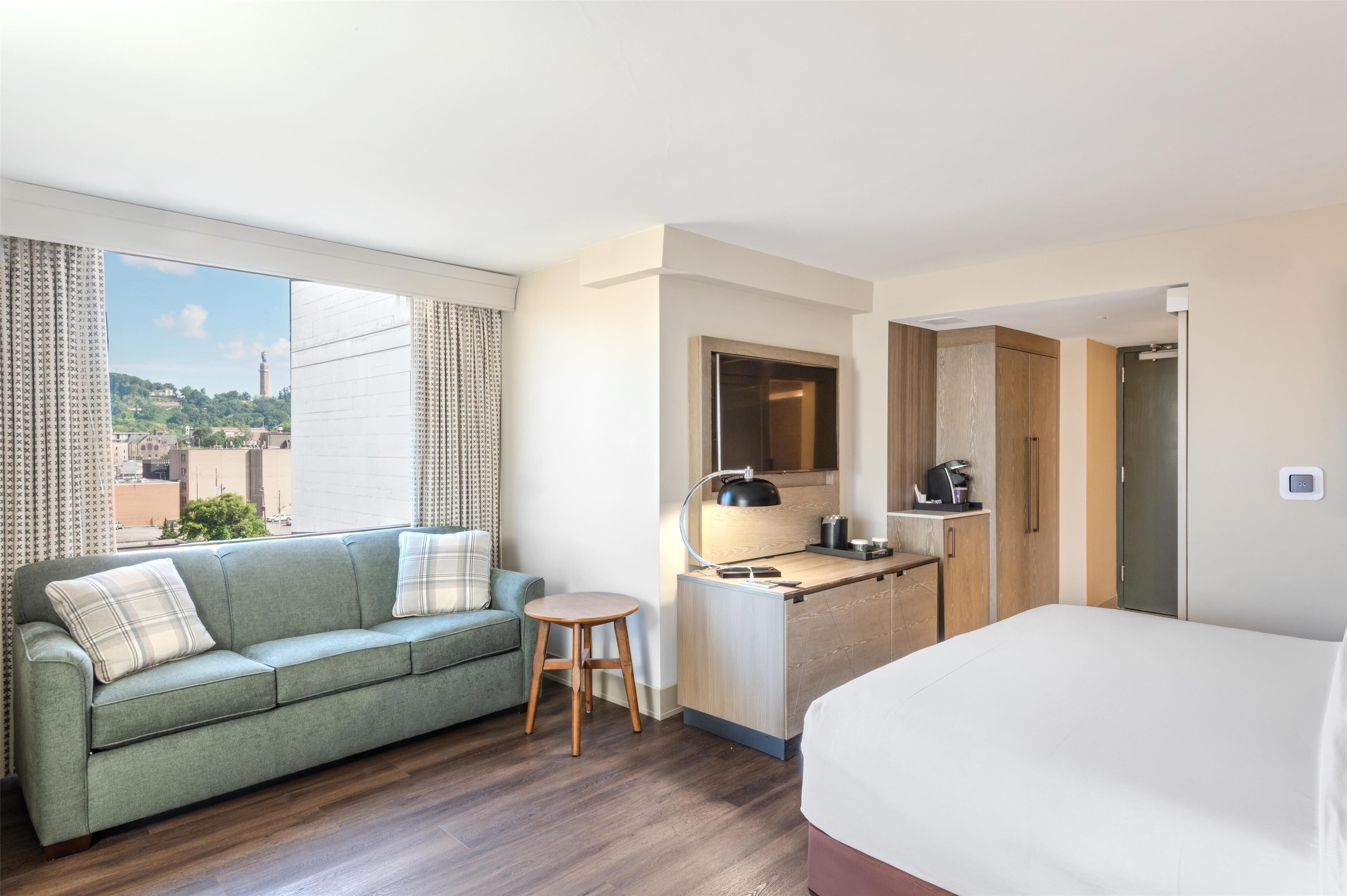 Habitación de hotel con sofá, un taburete pequeño, cama, kitchenette y vistas a la ciudad.