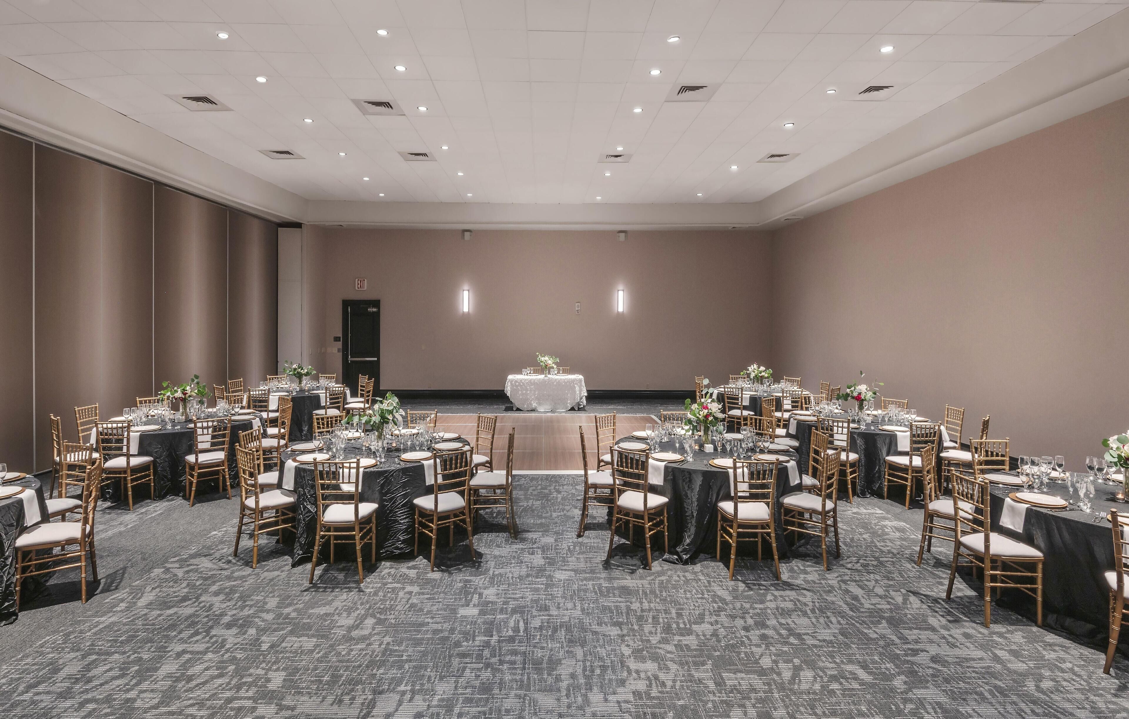 A large conference room with multiple tables and chairs, carpeted floor, and a stage at the front.