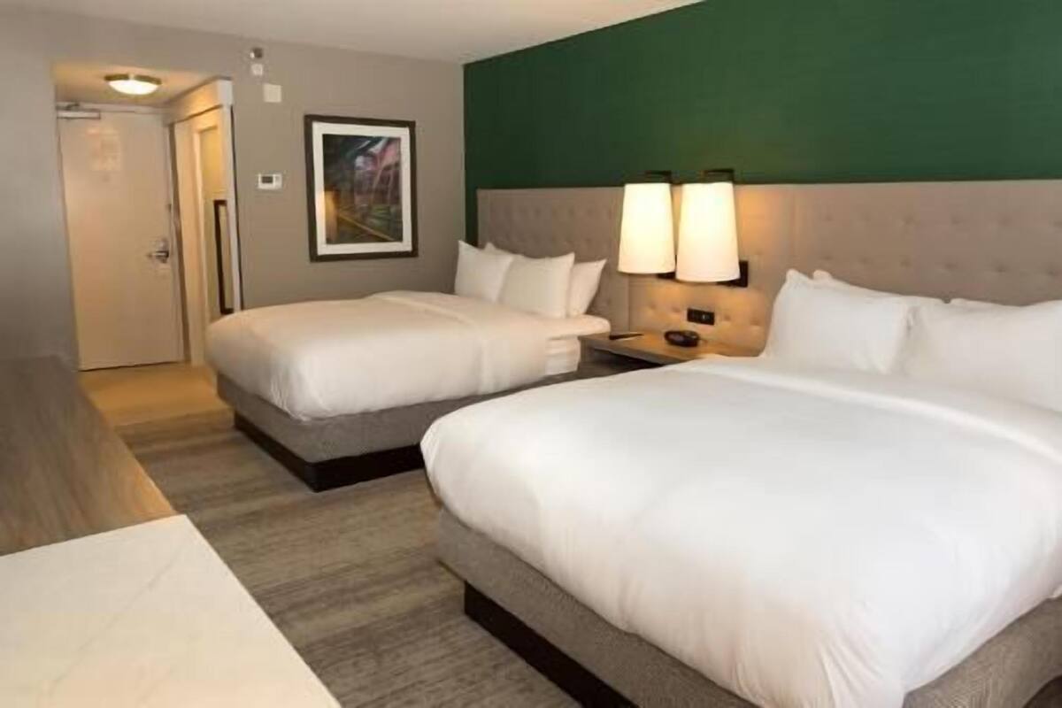 A hotel room with two beds, a nightstand with lamps, a framed picture on the wall, and a door in the background.