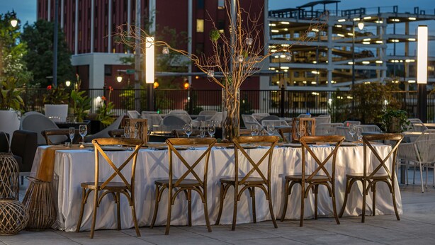 An outdoor dining area with tables set for six, surrounded by wicker chairs and a decorative tree centerpiece.