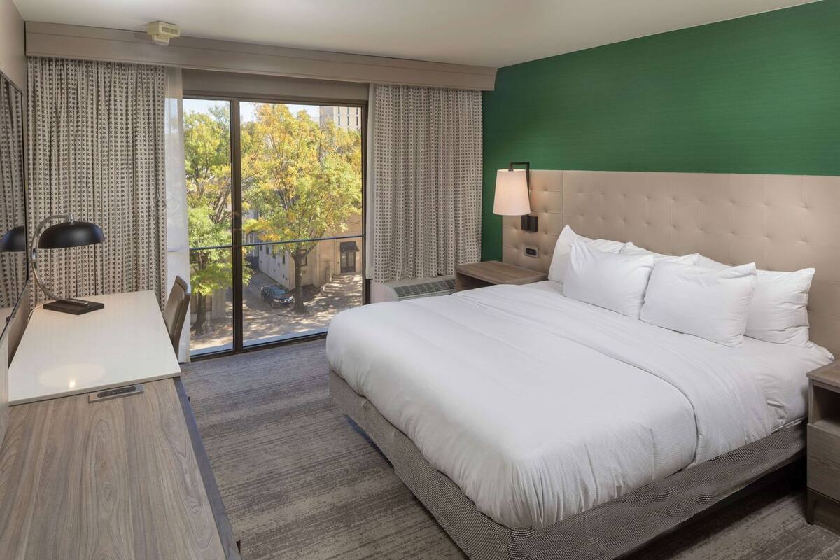 A hotel room with a bed, a desk, a chair, a lamp, and a view of the outdoors.