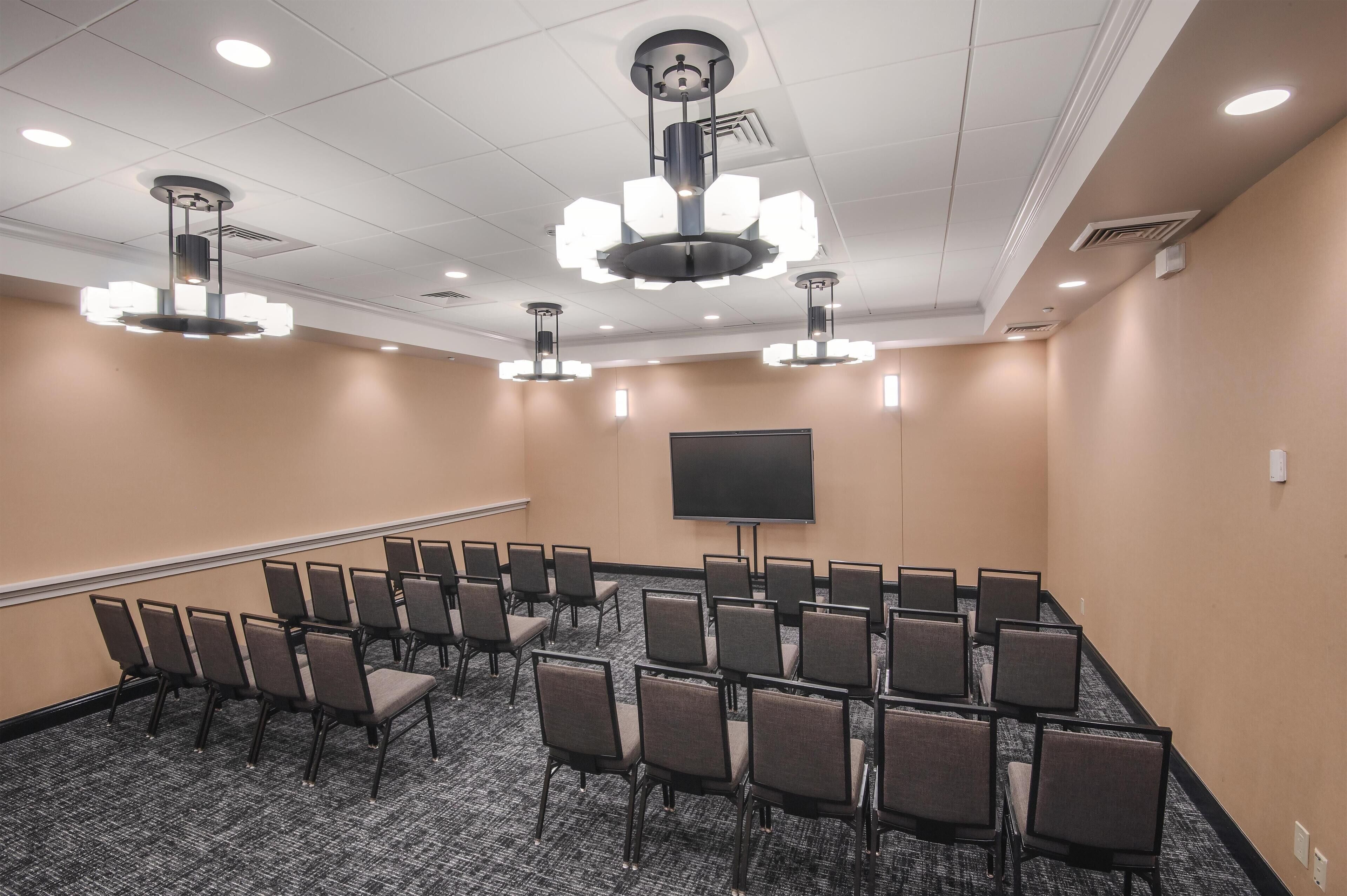 A conference room with rows of chairs, a large screen, and multiple ceiling lights.